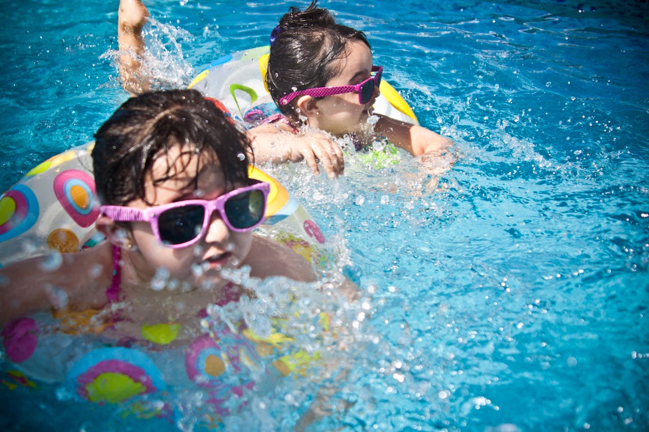 services-03 Two young girls enjoying a playful day in a bright blue swimming pool with colorful float rings.